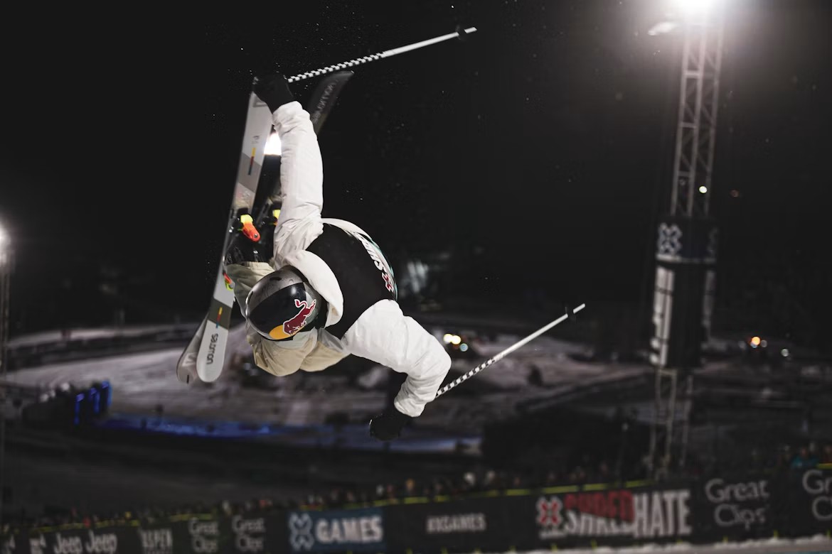 A freestyle skier performing an aerial trick under floodlights at night.